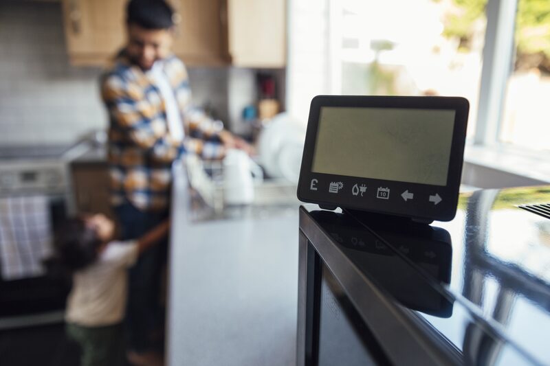 A smart meter in-home display on a kitchen counter with a blurred man and child in the background