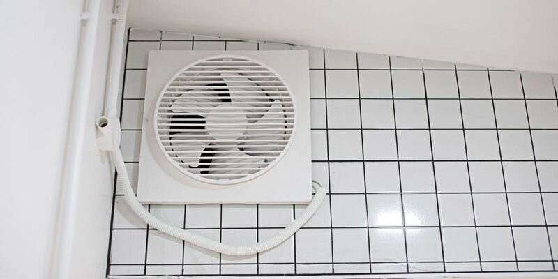 A white wall-mounted bathroom extractor fan with flexible ducting installed on a black-and-white tiled wall