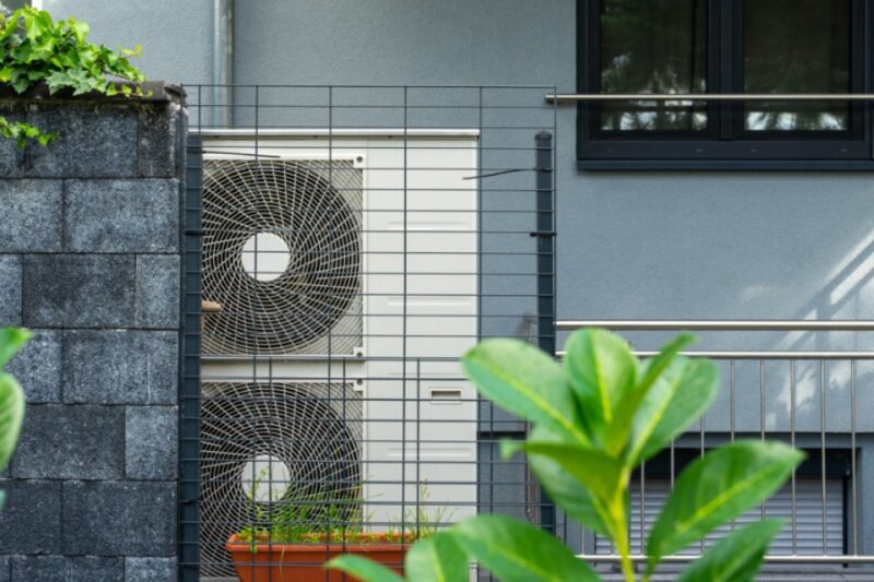 An air source heat pump unit with two fans mounted outside a grey building behind a metal railing