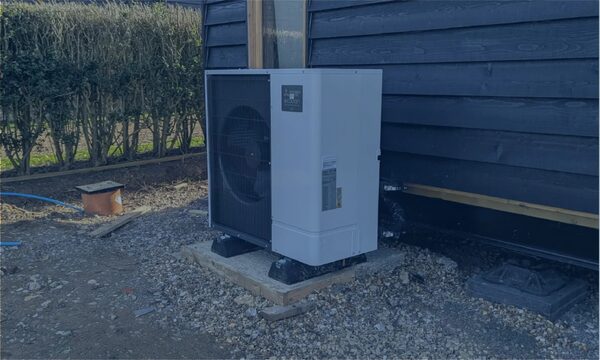 An air source heat pump unit installed outside a dark timber-clad building on a gravel base