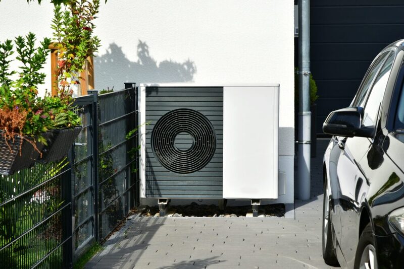 An air source heat pump unit installed on the outside wall of a house beside a driveway with a parked car