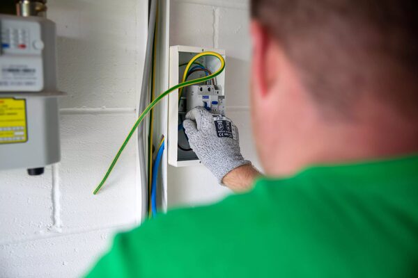 An engineer in a green shirt and gloves wiring an electrical consumer unit next to a gas meter on a white brick wall