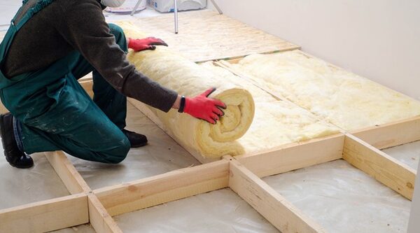 A worker in green overalls and red gloves unrolling a roll of yellow mineral wool loft insulation between wooden joists