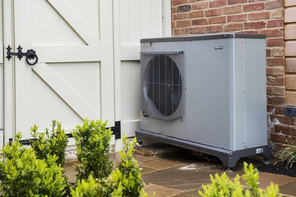A grey air source heat pump unit installed outside a brick house next to cream garage doors