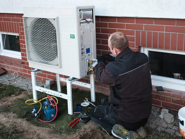 A technician installing an outdoor air source heat pump unit on a frame against a brick house wall