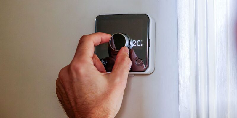 A hand turning the dial on a wall-mounted smart thermostat set to 20 degrees