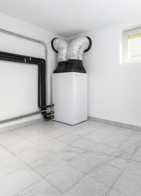 A white indoor heat pump or ventilation unit installed in a corner of a tiled utility room, with insulated flexible ducts rising from the top and pipework along the wall