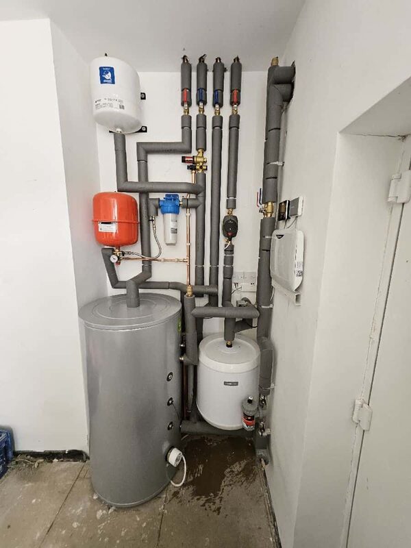 A heat pump plant room with a large grey cylinder, expansion vessels, filter and insulated pipework installed in a corner of a house