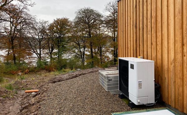 An air source heat pump unit installed outside a timber-clad house beside a gravel path with trees and a loch in the background