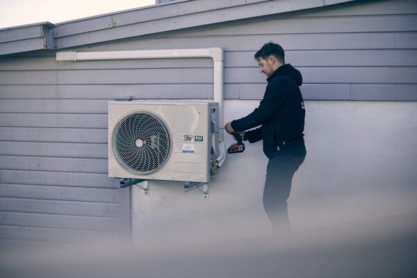 A technician using a power drill to install a wall-mounted air source heat pump unit on the exterior of a house