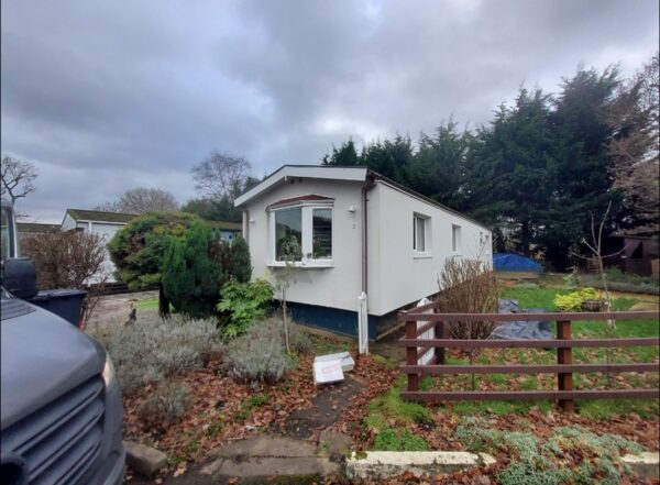 A white park home (mobile home) on a residential park, with a van partially visible in the foreground and a fenced garden