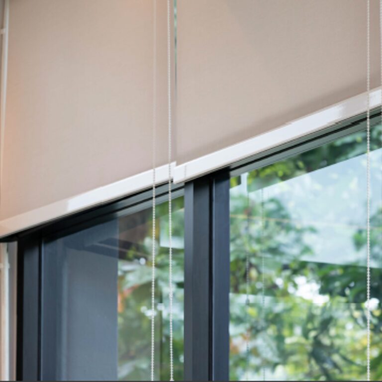 A beige roller blind partly lowered over a large window with greenery visible outside