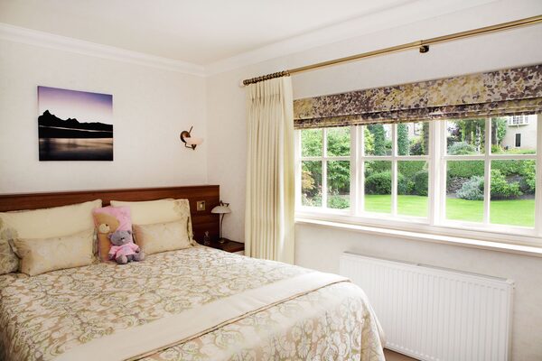 A bright bedroom with a large multi-paned sash window, curtains, a radiator beneath it and a made bed