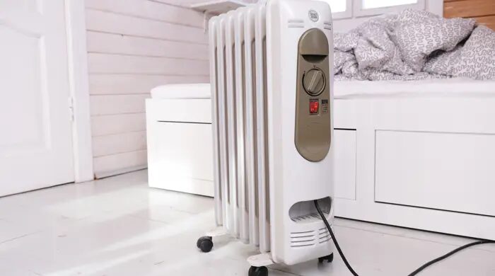 A white portable oil-filled radiator heater plugged in on the floor of a bright bedroom