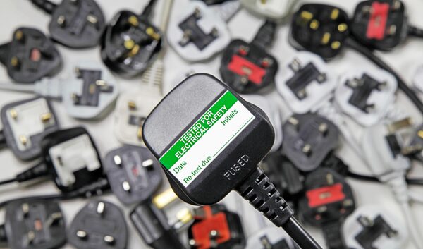 A close-up of a UK three-pin plug with a 'Tested for Electrical Safety' PAT testing label, surrounded by a pile of other plugs
