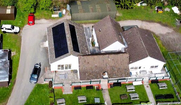 Aerial view of a large house with solar panels on its roof, surrounded by gardens and parked cars