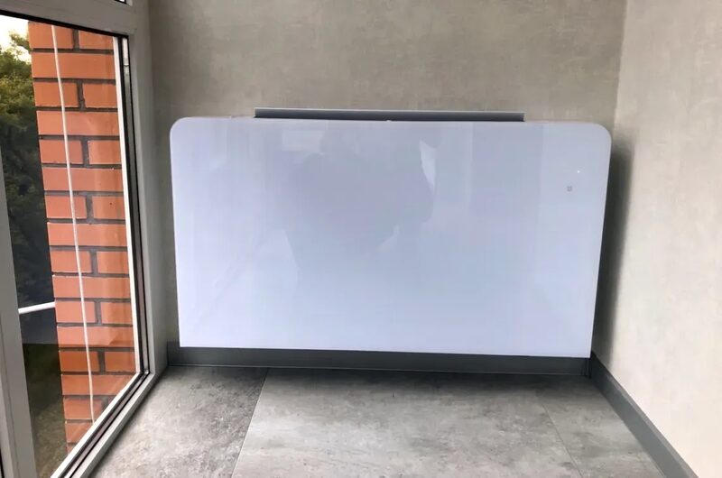 A white glass-panel electric heater installed on the wall of an apartment balcony corner next to a window