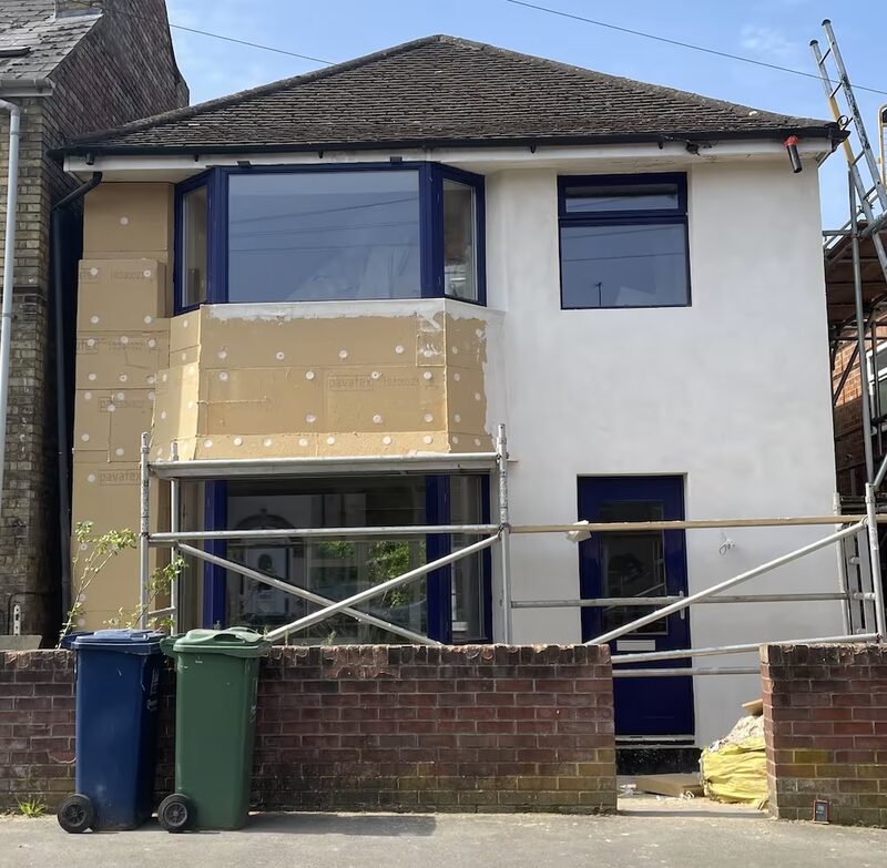 A house undergoing external solid wall insulation, with yellow insulation boards fixed to the front wall and scaffolding around it