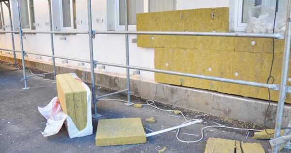 Yellow mineral wool insulation slabs being fitted to the outside wall of a building behind scaffolding, with offcuts and packaging on the ground