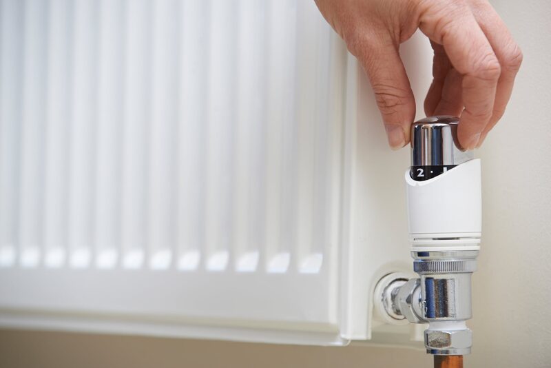 A hand turning the thermostatic radiator valve dial on a white panel radiator