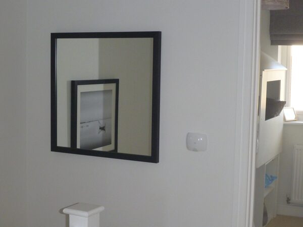 A small white smart-home sensor mounted on a wall next to a black-framed mirror in a house hallway