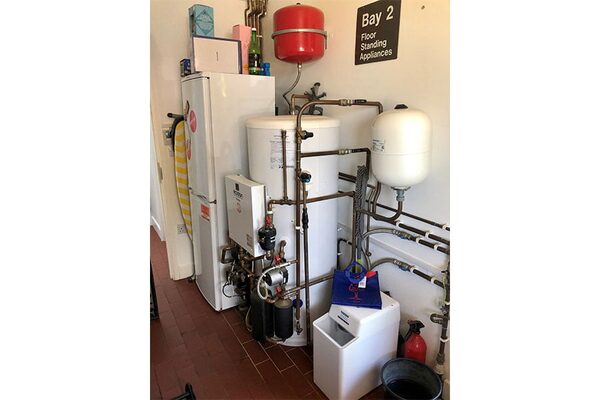 A cluttered utility room with a hot water cylinder, pipework, expansion vessels and a heat pump unit installed next to a fridge