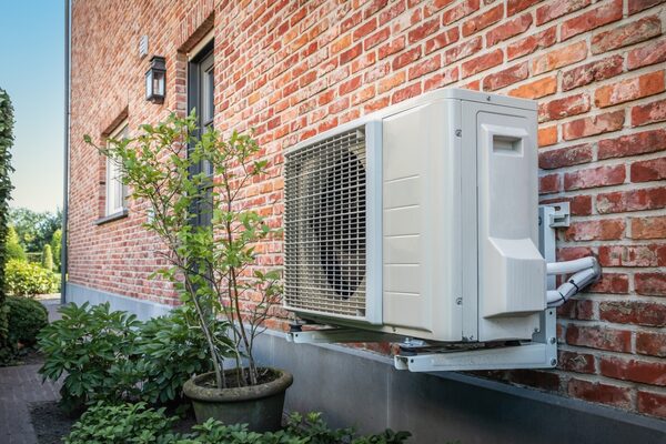 An air source heat pump unit mounted on the exterior brick wall of a house beside a potted shrub