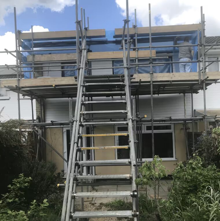 A house undergoing retrofit works, wrapped in scaffolding with a worker on the upper level and blue sheeting over the roof