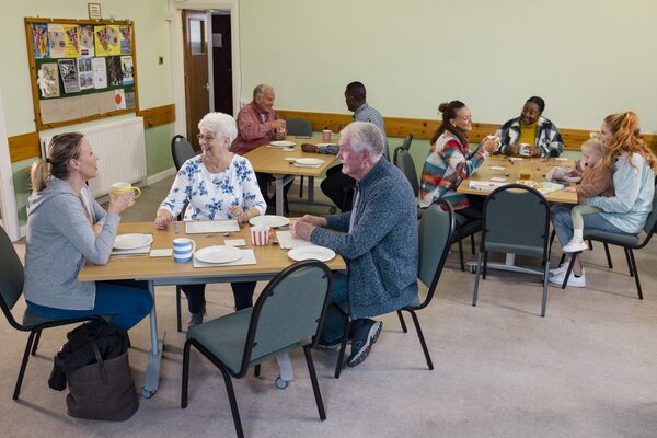 Diverse people of various ages chatting over coffee and food at tables in a community hall