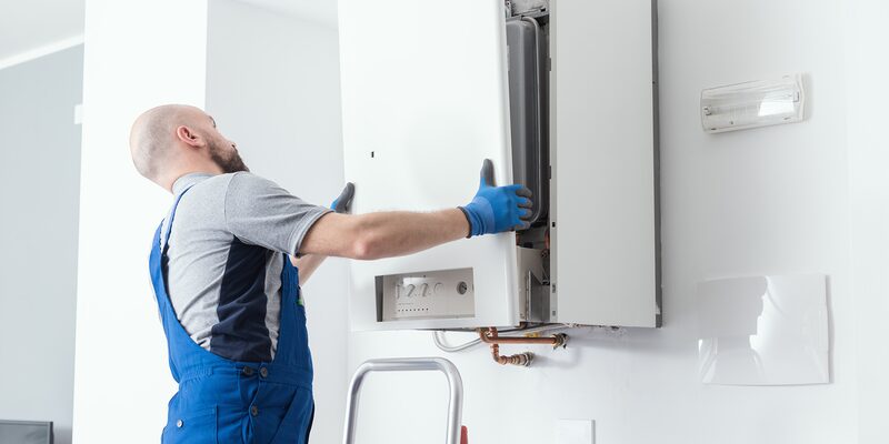 An engineer in blue overalls and gloves servicing a wall-mounted gas boiler with the casing open