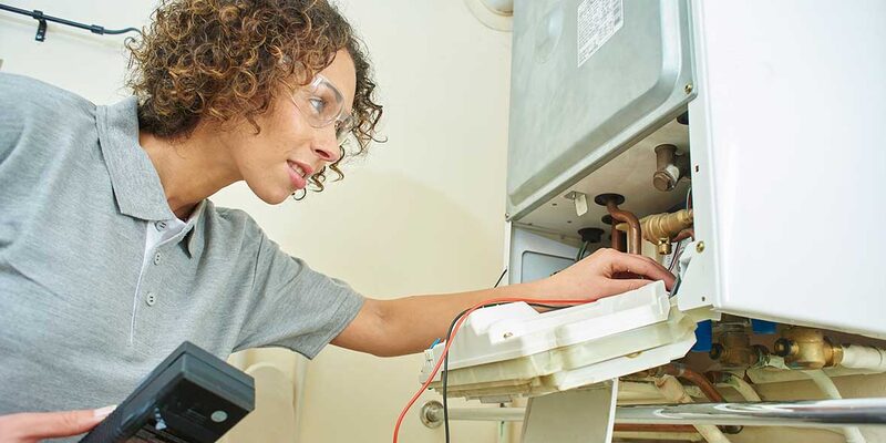 A technician with safety glasses inspecting the open internals of a wall-mounted gas boiler while holding a multimeter