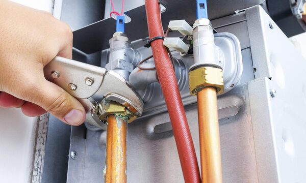 A hand using an adjustable wrench on a copper pipe fitting connected to valves on a boiler's metal casing