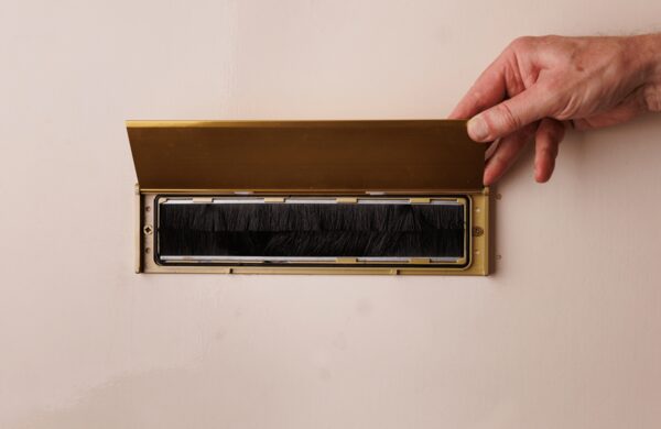 A hand lifting the brass flap of a letterbox fitted with a black brush draught excluder in a pale pink door