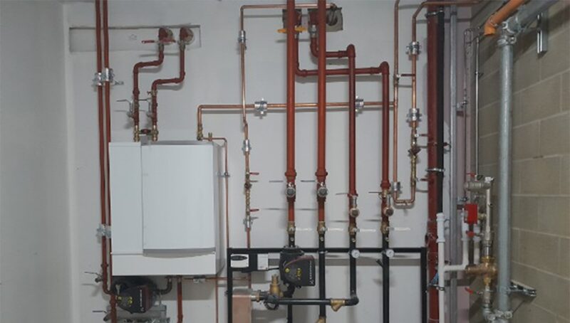 A white wall-mounted heating unit connected to an array of red and copper pipework, valves and meters in a plant room