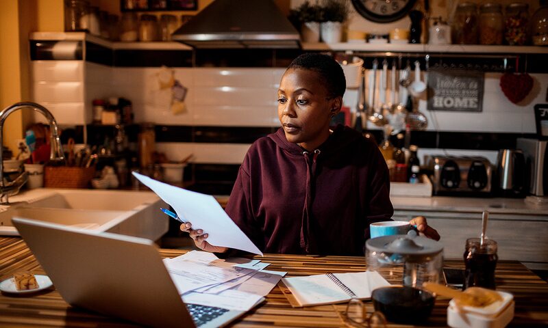 A woman sitting at a kitchen table at night reviewing a paper energy bill next to a laptop
