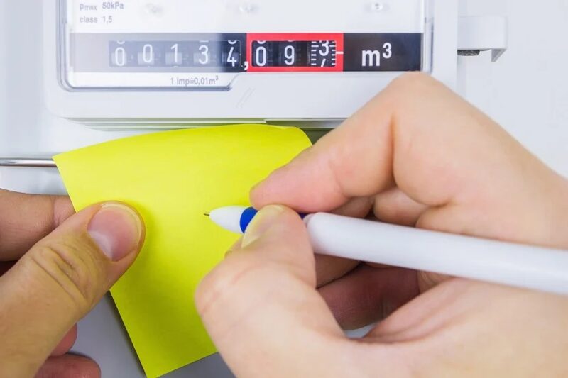 Hands writing on a yellow sticky note in front of a gas meter dial display reading in cubic metres