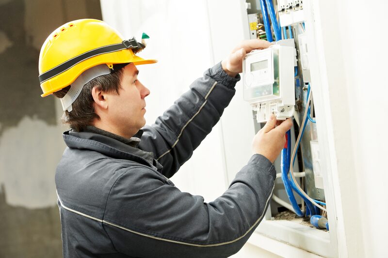 An engineer in a yellow hard hat installing a smart electricity meter in a wall-mounted consumer unit