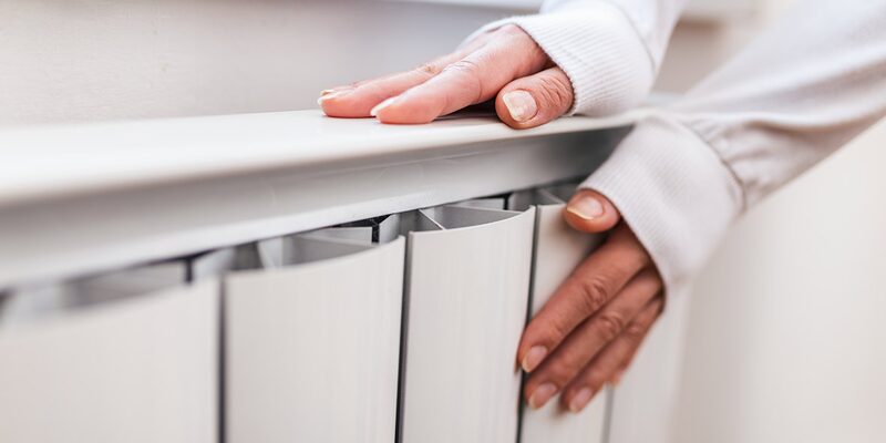 A person's hands resting on and feeling the warmth of a white panel radiator