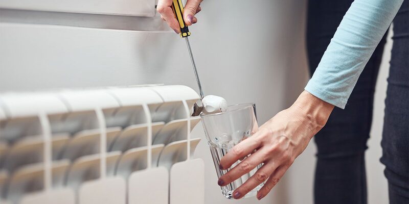 A person using a radiator key to bleed a white radiator while holding a glass underneath to catch water