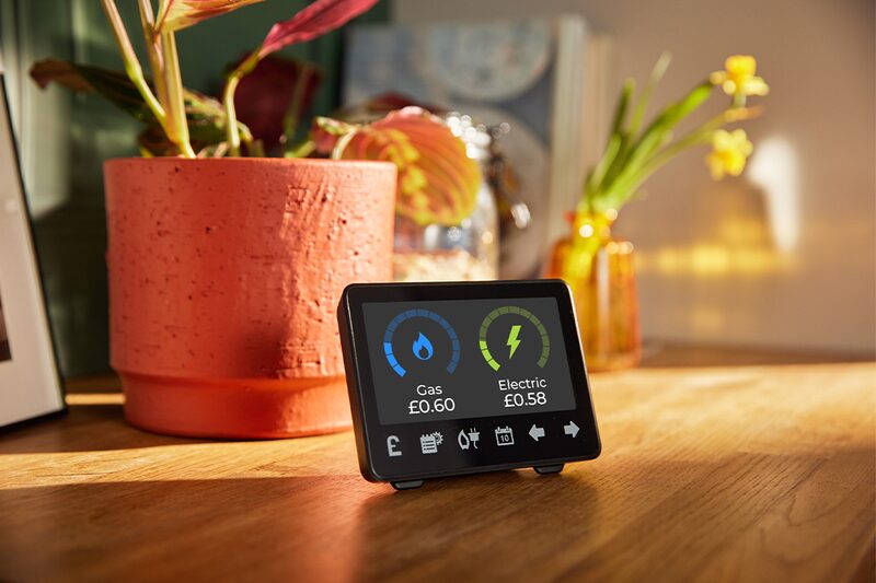 A smart meter display on a wooden table showing gas and electricity costs, with houseplants in the background