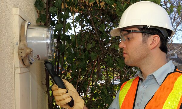 A technician in a hard hat and hi-vis vest reading an outdoor electricity meter on a wall with a handheld device