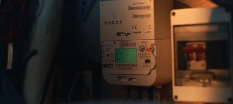 A Toshiba smart electricity meter with a green display mounted on a wall next to a consumer unit in dim lighting