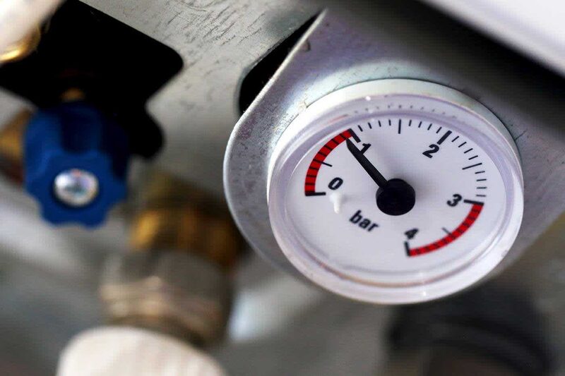 A close-up of a boiler pressure gauge reading about 1 bar, mounted on a boiler with a blue filling valve nearby