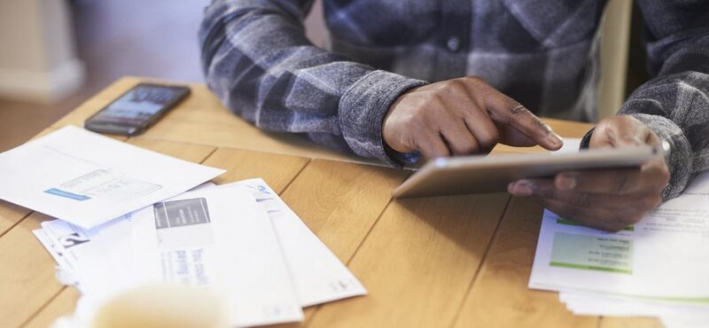 A person at a wooden table using a tablet beside scattered letters and a smartphone, reviewing household bills