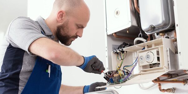 An engineer in blue overalls and gloves repairing the internal components of a wall-mounted gas boiler