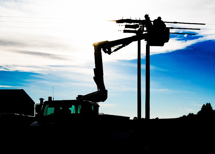 Silhouetted workers in a cherry picker on a cherry picker truck working on overhead electricity lines and poles against a bright sky