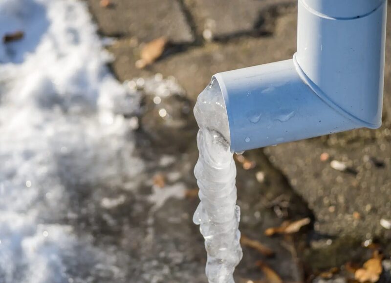 A white plastic condensate pipe with a column of ice frozen at its open end, outdoors on snowy ground