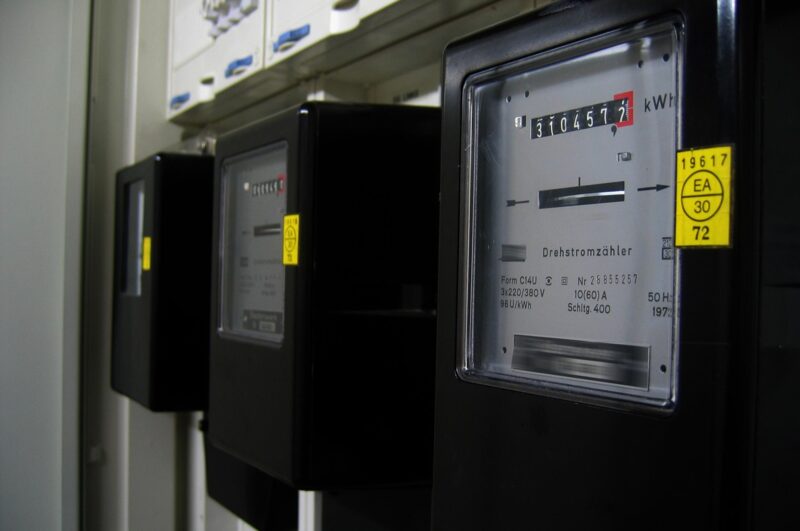 A row of black electricity meters mounted on a wall, with the nearest meter's dial face and yellow calibration sticker in focus