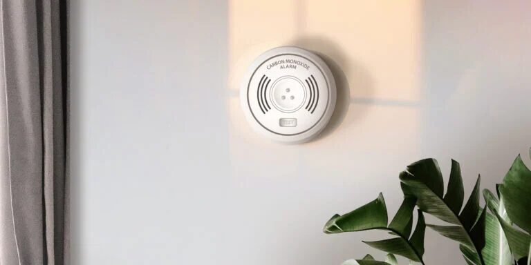 A white carbon monoxide alarm mounted on a wall next to a curtain and a houseplant