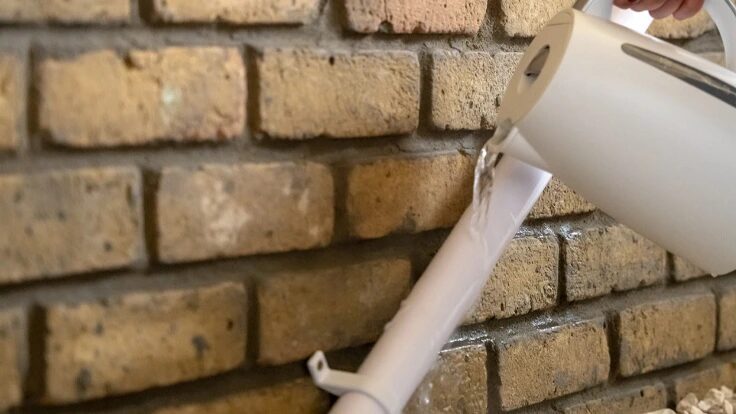 A hand pouring warm water from a white kettle over a white condensate pipe on a brick wall to thaw it